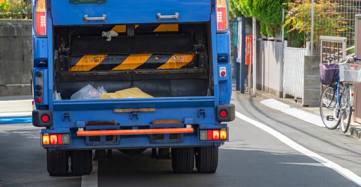 Caminhão de lixo azul realizando coleta urbana em via residencial durante o dia