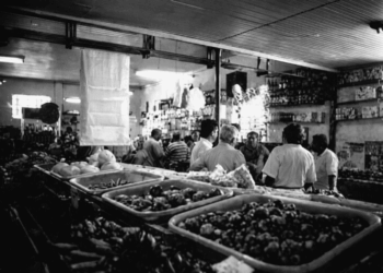 Interior do Bar Cinédia em Itabira com clientes reunidos ao balcão, prateleiras com bebidas ao fundo e bandejas de alimentos em primeiro plano, local histórico ligado à língua do camaco