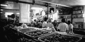 Interior do Bar Cinédia em Itabira com clientes reunidos ao balcão, prateleiras com bebidas ao fundo e bandejas de alimentos em primeiro plano, local histórico ligado à língua do camaco