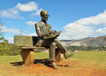 Estátua de Carlos Drummond de Andrade sentada em banco ao ar livre, em Itabira, com montanhas ao fundo.