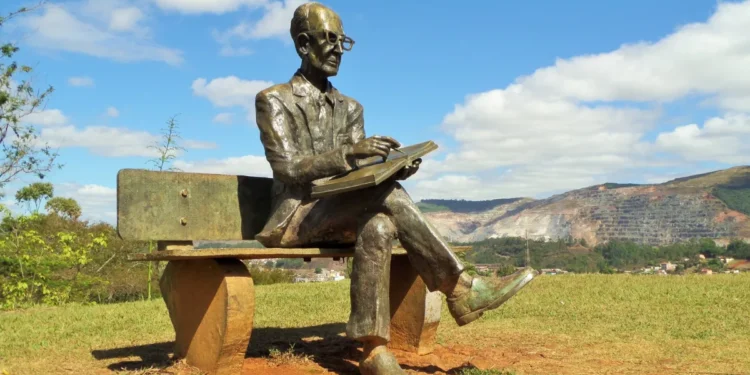 Estátua de Carlos Drummond de Andrade sentada em banco ao ar livre, em Itabira, com montanhas ao fundo.