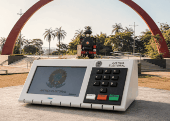Urna eletrônica em destaque na Praça do Areão em Itabira com a Maria Fumaça ao fundo, representando as eleições na cidade