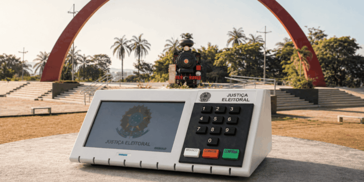 Urna eletrônica em destaque na Praça do Areão em Itabira com a Maria Fumaça ao fundo, representando as eleições na cidade