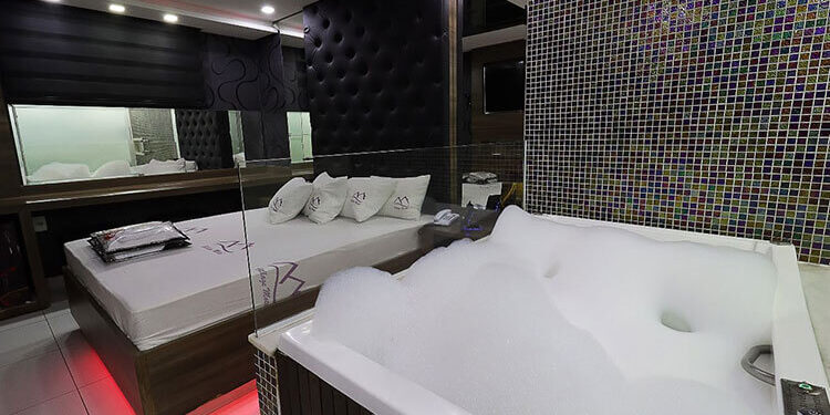 Suíte de motel em Itabira com cama, espelhos, iluminação moderna e banheira de hidromassagem com espuma.
